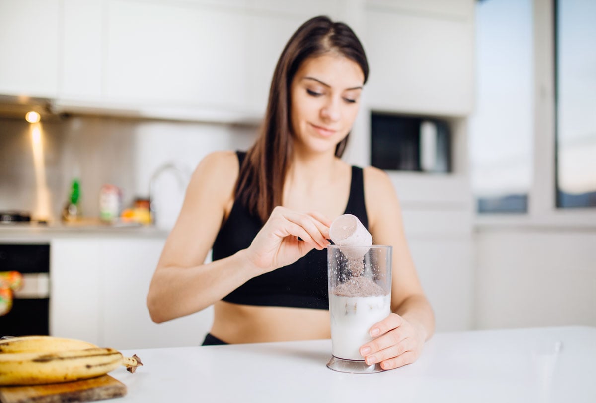 Woman in sports attire pours chocolate protein powder into glass in kitchen.